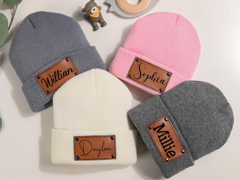 K&ouml;nnte beinhalten: Eine Auswahl an Baby-M&uuml;tzen in Grau, Rosa, Creme und Dunkelgrau. Jede M&uuml;tze hat ein braunes Lederpatch mit einem Namen in schwarzer Schrift. Die Namen sind Willian, Sophia, Daylan und Millie. Ein sch&ouml;nes Geschenk.