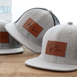 May include: Three gray baseball caps with personalized leather patches. Each cap has a different name in cursive script: Jardin, Heber, and Petra. The caps have a flat brim and a mesh back.