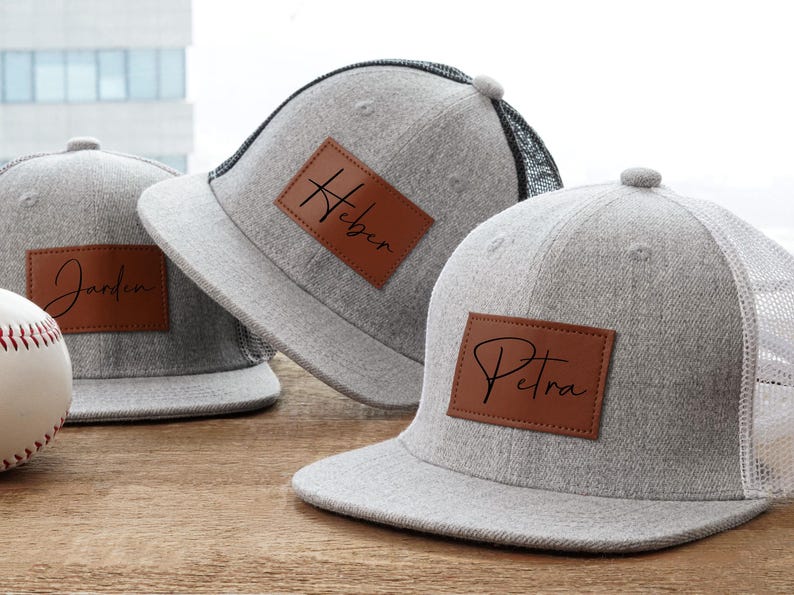 May include: Three gray baseball caps with personalized leather patches. Each cap has a different name in cursive script: Jardin, Heber, and Petra. The caps have a flat brim and a mesh back.