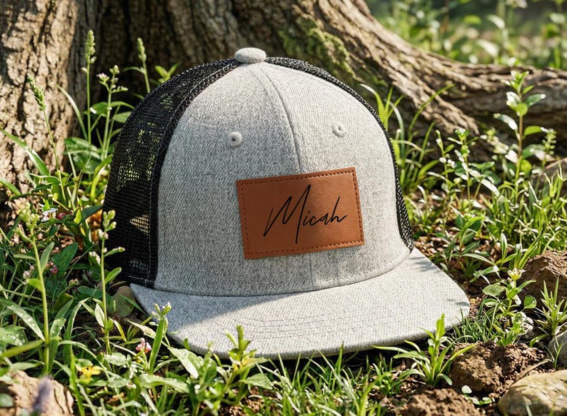 May include: A gray trucker hat with a black mesh back and a curved brim. The hat features a brown leather patch with the name "Micah" in a handwritten style. The hat is sitting in grass.