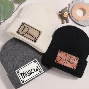 K&ouml;nnte beinhalten: Drei Strickm&uuml;tzen in Creme, Grau und Schwarz, jede mit einem rechteckigen Aufn&auml;her mit einem Namen. Die cremefarbene M&uuml;tze hat "Dennis" in Schrift, die graue "Marcus" und die schwarze "Albert". Kleine dekorative Spielzeuge sind im Hintergrund.