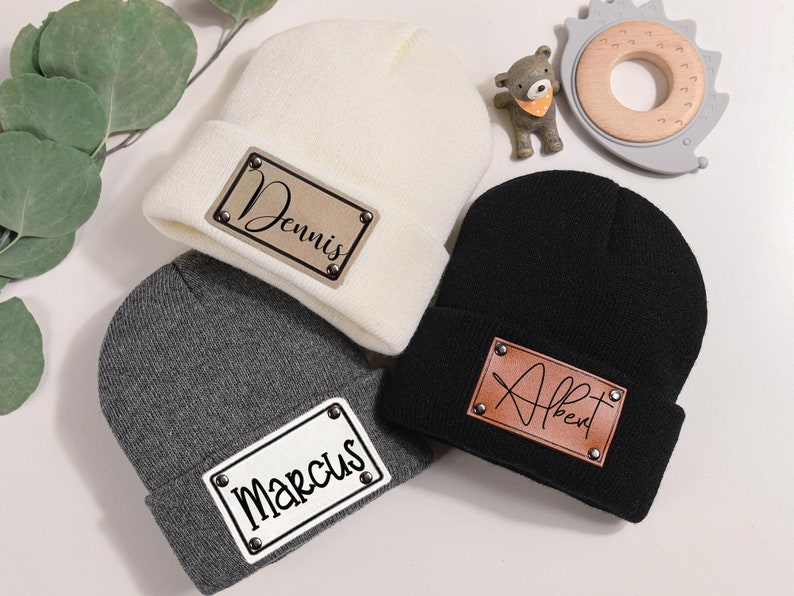 K&ouml;nnte beinhalten: Drei Strickm&uuml;tzen in Creme, Grau und Schwarz, jede mit einem rechteckigen Aufn&auml;her mit einem Namen. Die cremefarbene M&uuml;tze hat "Dennis" in Schrift, die graue "Marcus" und die schwarze "Albert". Kleine dekorative Spielzeuge sind im Hintergrund.