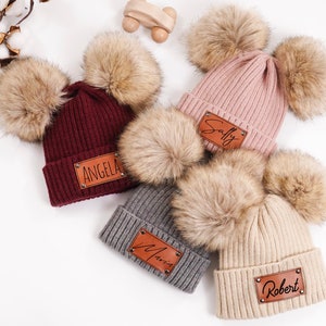 May include: A collection of ribbed knit beanies in various colors, each adorned with two large faux fur pom-poms. Each beanie features a rectangular leather patch with a name on it. Colors include burgundy, pink, gray, and beige.