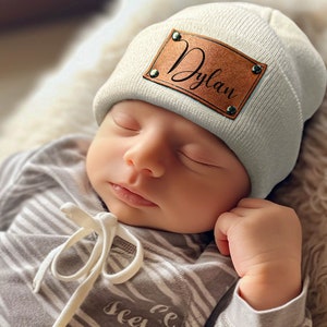 K&ouml;nnte beinhalten: Ein schlafendes Baby tr&auml;gt eine hellgraue M&uuml;tze mit einem braunen Lederaufn&auml;her mit dem Schriftzug "Dylan" in Schreibschrift. Das Baby tr&auml;gt au&szlig;erdem einen grau-wei&szlig; gestreiften Strampler.