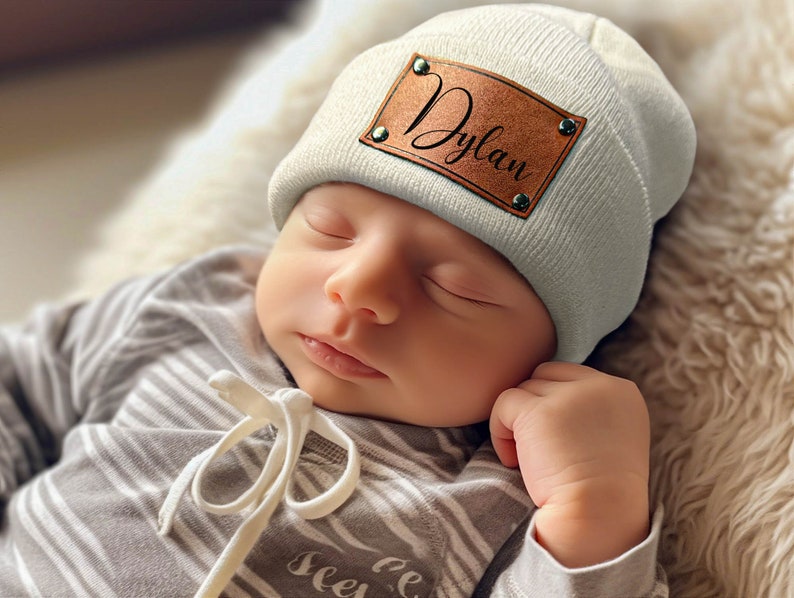 K&ouml;nnte beinhalten: Ein schlafendes Baby tr&auml;gt eine hellgraue M&uuml;tze mit einem braunen Lederaufn&auml;her mit dem Schriftzug "Dylan" in Schreibschrift. Das Baby tr&auml;gt au&szlig;erdem einen grau-wei&szlig; gestreiften Strampler.
