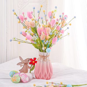 May include: A bouquet of pastel-colored artificial flowers in a pink vase. The arrangement includes pink tulips, small white flowers, and colorful spherical accents. Easter eggs and a wooden bunny decoration are also present.