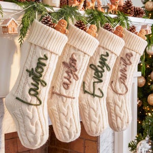 May include: Four cream-coloured cable knit Christmas stockings hanging in a row. Each stocking features a folded cuff, a small brown pinecone, and a gingerbread man figure at the top. The names Layton, Taylor, Evelyn, and Camila are embroidered.
