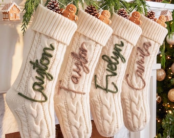 Op de afbeelding: Vier crèmekleurige kerstsokken van kabelbreisel die op een rij hangen. Elke sok heeft een omgeslagen boord, een kleine bruine dennenappel en een peperkoekmannetje aan de bovenkant. De namen Layton, Taylor, Evelyn en Camila zijn geborduurd.
