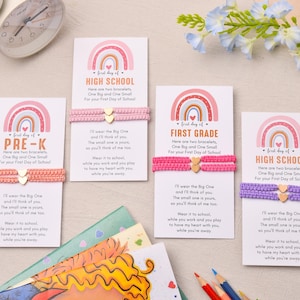 May include: Four white rectangular cards featuring a rainbow graphic and text for Pre-K, First Grade, and High School. Each card holds two woven bracelets in coral, pink, or purple, each featuring a small gold-tone heart charm in the center.