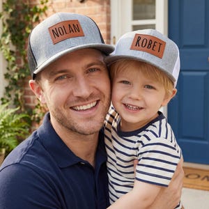 May include: Two gray trucker hats with leather patches. One hat reads "NOLAN" and the other reads "ROBET". The hats are worn by a man and a child, both smiling. The man wears a navy blue polo shirt, and the child wears a striped shirt.