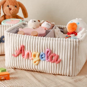 Può includere: Cesto portaoggetti in velluto a coste color crema con il nome "Angela" in lettere di filo colorate. Il cesto è pieno di giocattoli, tra cui un'anatra di peluche, un'auto di legno rosa e un peluche. Un secondo cesto con il nome "Wyatt" è visibile.