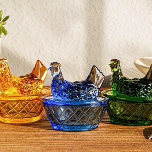 Puede incluir: Tres recipientes de vidrio con forma de gallina en ámbar, azul y verde. Cada gallina se asienta sobre una base en forma de cesta. Los recipientes están sobre una superficie de madera, con una planta en una maceta blanca al fondo.