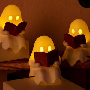 Puede incluir: Tres pequeñas figuras de fantasmas de color blanco roto, cada una emitiendo un cálido resplandor amarillo desde sus ojos. Cada fantasma está sentado y sostiene un libro abierto de color rojo oscuro en sus manos.