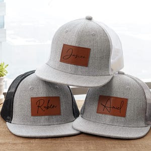 May include: Three gray trucker hats with personalized brown leather patches. Each hat has a different name: Jasira, Ruben, and Ariel. The hats have a mesh back and a flat brim.