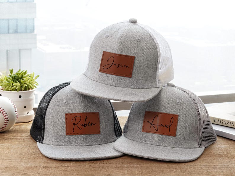 May include: Three gray trucker hats with personalized brown leather patches. Each hat has a different name: Jasira, Ruben, and Ariel. The hats have a mesh back and a flat brim.