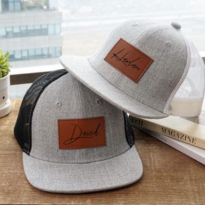 May include: Two gray baseball caps with personalized leather patches. One cap has a black mesh back and the name "David" on the patch. The other has a white mesh back and the name "Harlan". Both caps are on a wooden surface.