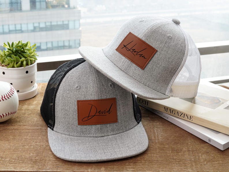 May include: Two gray baseball caps with personalized leather patches. One cap has a black mesh back and the name "David" on the patch. The other has a white mesh back and the name "Harlan". Both caps are on a wooden surface.