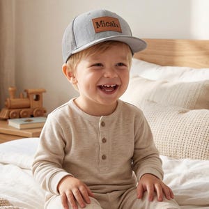 May include: A gray trucker hat with a brown leather patch that reads "Micah". The hat is worn by a child. The child is wearing a beige long-sleeved shirt and is sitting on a bed with white bedding.