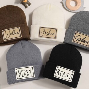 K&ouml;nnte beinhalten: F&uuml;nf Strickm&uuml;tzen in verschiedenen Farben: Braun, Creme, Grau und Schwarz. Jede M&uuml;tze hat ein rechteckiges Patch mit einem Namen: Walker, Joshua, Robert, Herry und Remy. Die Patches haben einen hellbraunen Hintergrund mit schwarzer Schrift.