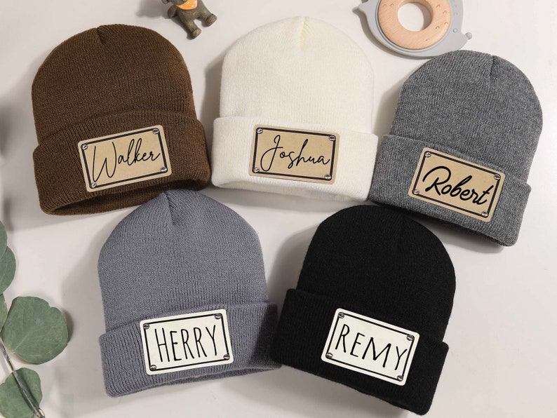 K&ouml;nnte beinhalten: F&uuml;nf Strickm&uuml;tzen in verschiedenen Farben: Braun, Creme, Grau und Schwarz. Jede M&uuml;tze hat ein rechteckiges Patch mit einem Namen: Walker, Joshua, Robert, Herry und Remy. Die Patches haben einen hellbraunen Hintergrund mit schwarzer Schrift.