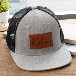 May include: Gray trucker hat with a black mesh back and a brown leather patch. The patch features the name "Kohen" in a handwritten style. The hat is sitting on a wooden surface, with a baseball and a small plant in the background.