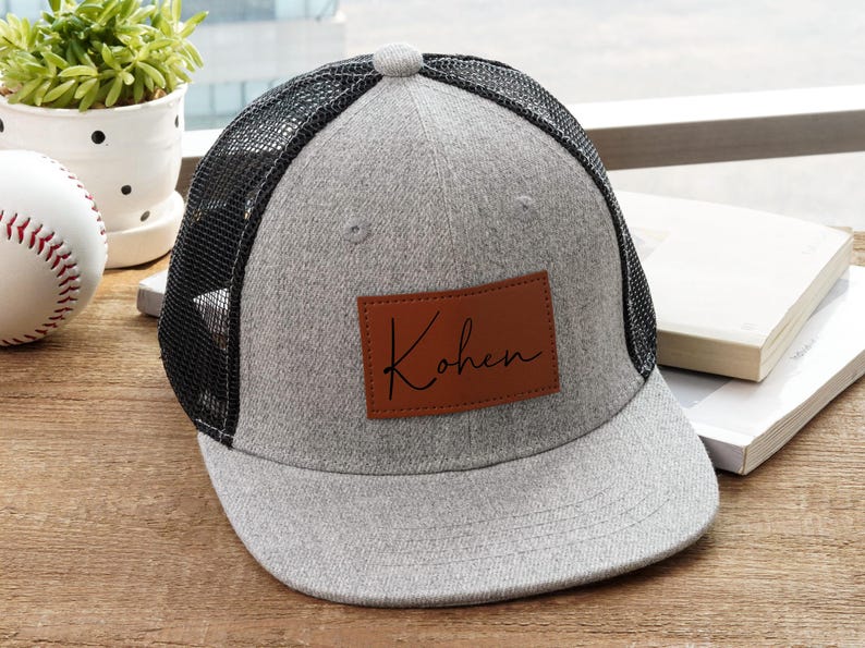 May include: Gray trucker hat with a black mesh back and a brown leather patch. The patch features the name "Kohen" in a handwritten style. The hat is sitting on a wooden surface, with a baseball and a small plant in the background.