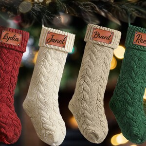 Peut inclure: Quatre bas de Noël en tricot torsadé, rouges, crème, beige et verts, chacun avec une étiquette en cuir portant un nom : "Lydia", "Janet", "Brant" et "Reed". Les bas sont suspendus, avec un arrière-plan flou de lumières et de verdure de Noël.