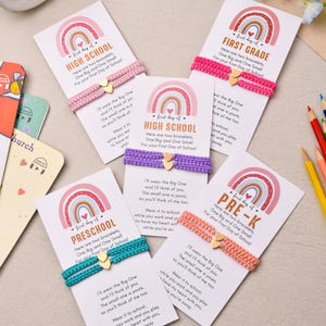 May include: Five sets of two woven bracelets on white cards, each featuring a small gold heart charm. The cards display a rainbow graphic and text including First Day of School, High School, Preschool, and Pre-K, with matching colored cords.