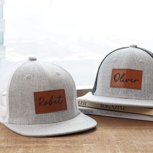 May include: Two gray trucker hats with personalized leather patches. One hat has "Robert" and the other "Oliver" in script. The hats have a gray crown and brim, with a white or black mesh back. A baseball sits nearby.