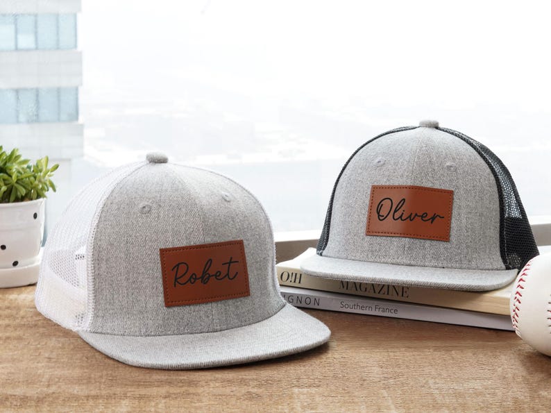 May include: Two gray trucker hats with personalized leather patches. One hat has "Robert" and the other "Oliver" in script. The hats have a gray crown and brim, with a white or black mesh back. A baseball sits nearby.