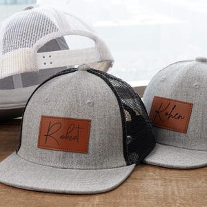 May include: Gray trucker hats with black mesh backs and brown leatherette patches. One hat has "Robert" and the other has "Kohen" in script. A white mesh-backed hat and a baseball are also in the image.