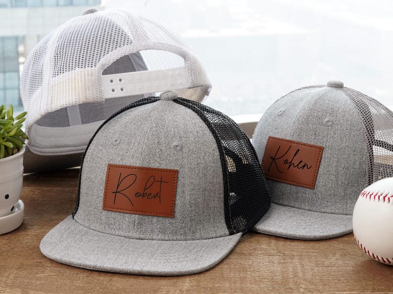 May include: Gray trucker hats with black mesh backs and brown leatherette patches. One hat has "Robert" and the other has "Kohen" in script. A white mesh-backed hat and a baseball are also in the image.