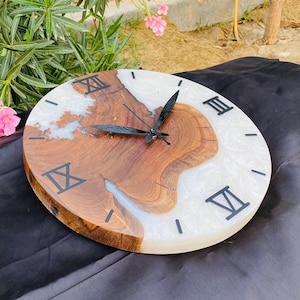 May include: A round wooden clock with a natural wood grain center and a white resin border. Black Roman numerals mark the hours. The clock hands are black. The clock is on a black surface.