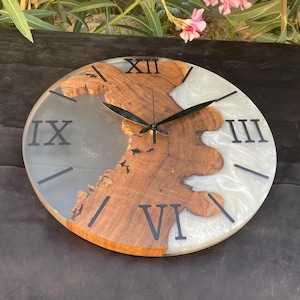 May include: Round wooden wall clock with a natural wood grain design, black Roman numerals, and black clock hands. The clock face has a section of gray resin and a section of white resin.