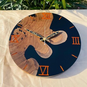 May include: Round wall clock with a unique design. The clock face features a combination of dark blue resin and natural wood grain. Roman numerals and clock hands are in a copper color. A decorative piece for any room.