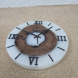 May include: White and brown round wall clock with Roman numeral markings. The clock face has a wood slice design in the center.