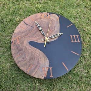 May include: A round wooden wall clock with a unique design. The clock face is split, with a dark blue section and a natural wood grain section. Roman numerals and clock hands are gold. The clock is on a green grassy surface.