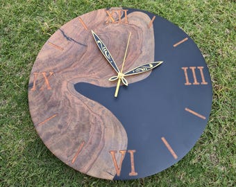 May include: A round wooden wall clock with a unique design. The clock face is split, with a dark blue section and a natural wood grain section. Roman numerals and clock hands are gold. The clock is on a green grassy surface.
