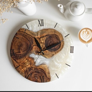 May include: A round wooden and white resin clock with Roman numeral markings. The clock has black hands and a white background with a wood grain pattern.