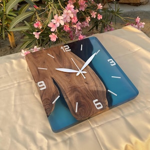 May include: Square wall clock with a wooden face and blue resin accents. The clock features white numerals and hands, with a natural wood grain pattern. The clock is approximately 10 inches square.
