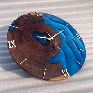 May include: A round wooden clock with a striking design. The clock face features a combination of dark brown wood and vibrant blue resin. Roman numerals mark the hours, and the clock hands are silver.