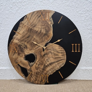 Puede incluir: Un reloj redondo de madera con resina epoxi negra. La esfera del reloj tiene números romanos y manecillas doradas.