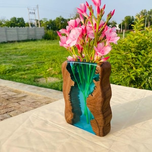 Pode incluir: Um vaso de madeira artesanal com um design único, apresentando um exterior de madeira natural e um interior de resina azul vibrante. O vaso contém um buquê de flores cor-de-rosa, criando um contraste marcante. O vaso tem cerca de 25 cm de altura.