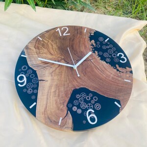 May include: Round wooden wall clock with a natural wood grain center and a dark blue resin border. White numerals mark the hours, and the clock hands are white. Decorative gears are embedded in the resin.
