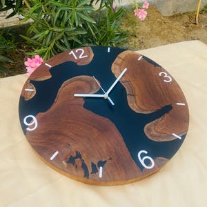 May include: Round wooden wall clock with a black epoxy resin center. The clock face features natural wood grain patterns, white numerals, and white clock hands. The clock is approximately 12 inches in diameter.