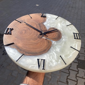 May include: A round wooden clock with a natural wood grain center and a white, swirled resin border. Black Roman numerals mark the hours, and black hands indicate the time. The clock is a decorative home accent.