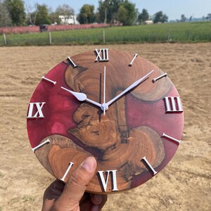 May include: A round wooden clock with a red resin background and natural wood grain patterns. White Roman numerals and clock hands provide contrast. The clock is held in a hand, showcasing its size and design.