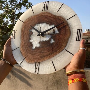 May include: White and brown resin clock with Roman numeral markings. The clock face is made of white resin with a brown wood slice embedded in the center. The clock hands are black.