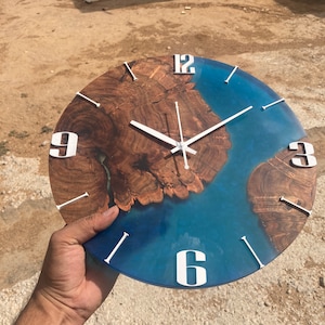 May include: Round wall clock with a unique design. The clock face features a combination of wood and blue resin, with white numerals and hands. The wood has a natural, brown grain pattern. The clock is a decorative item.