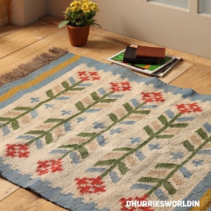 May include: A rectangular woven rug featuring a cream-colored base with a repeating pattern of red flowers and green stems. The rug has light blue borders with yellow stripes and fringed edges. The text DHURRIESWORLDIN is printed in the corner.